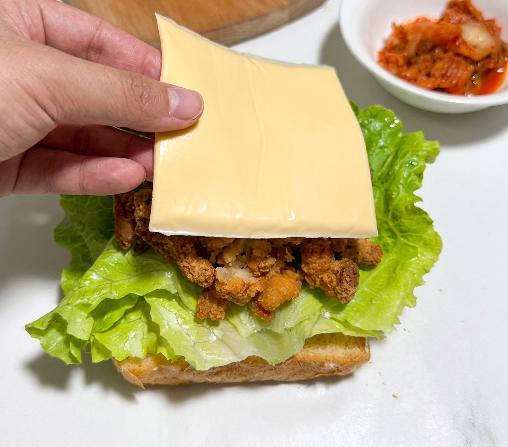 Une main plaçant une tranche carrée de fromage jaune sur des morceaux de poulet frit.