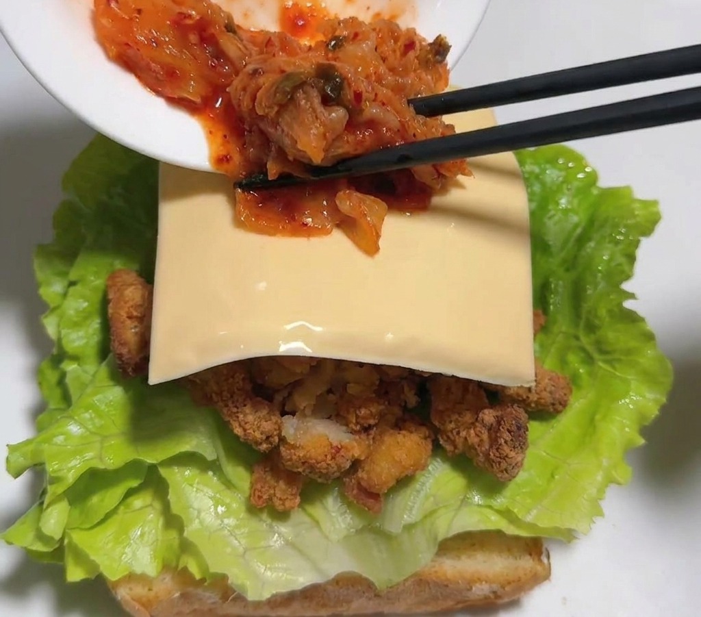 Des baguettes noires déposant du kimchi de chou rouge vif sur une tranche de fromage à lintérieur du sandwich.