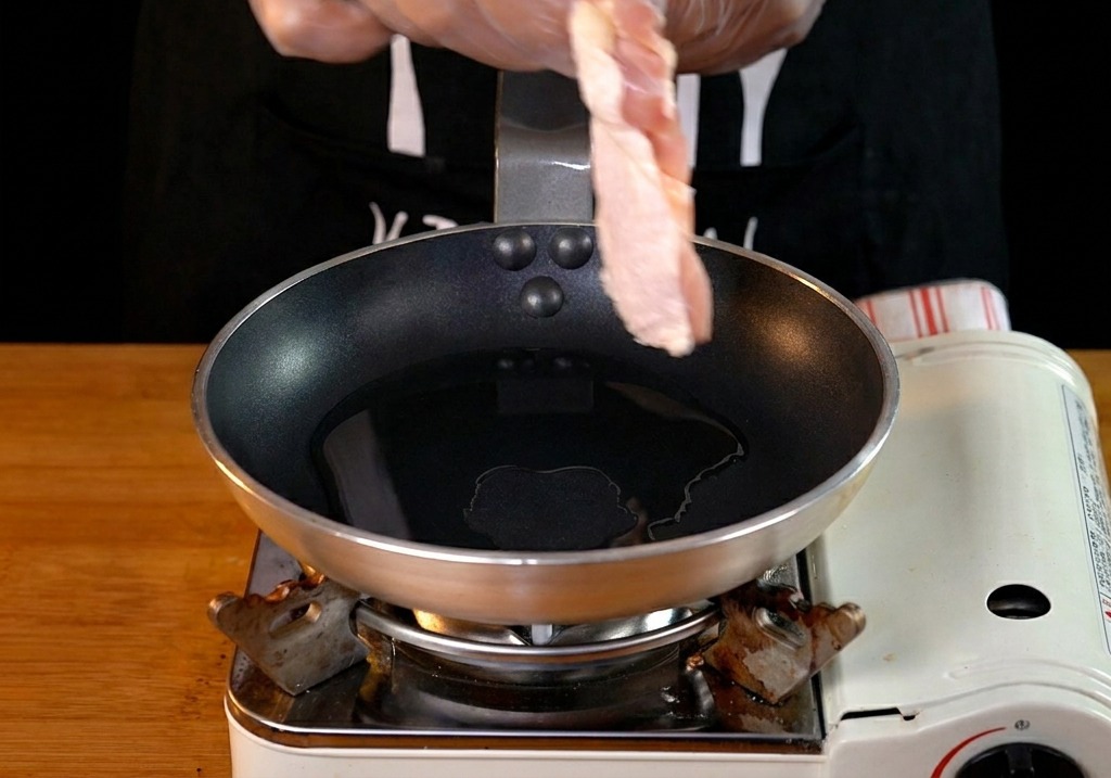 Une main plaçant un haut de cuisse de poulet cru dans une poêle avec de lhuile chaude.