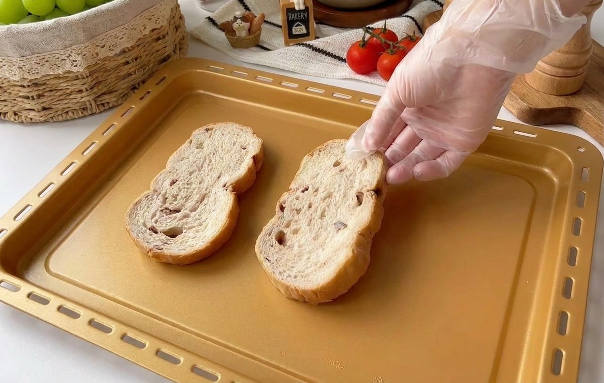 Une main gantée déposant deux tranches épaisses de pain multicéréales rustique sur une plaque de cuisson dorée.