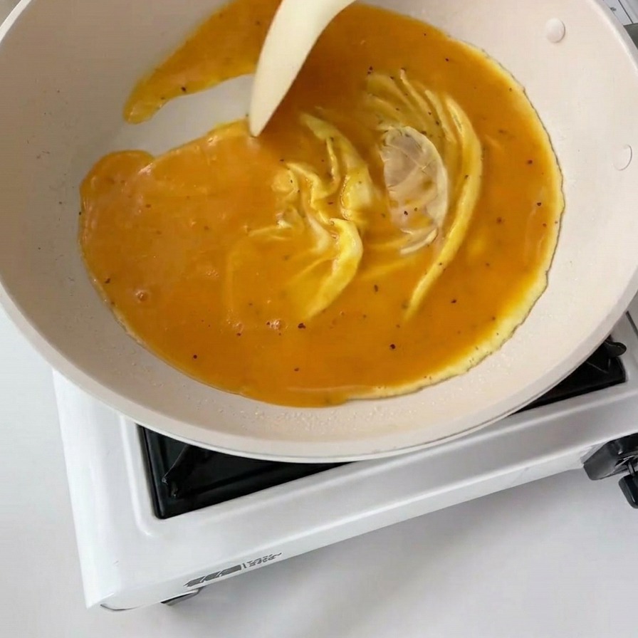 Une spatule blanche poussant les œufs partiellement cuits dans une poêle blanche, créant de larges plis jaunes onctueux.