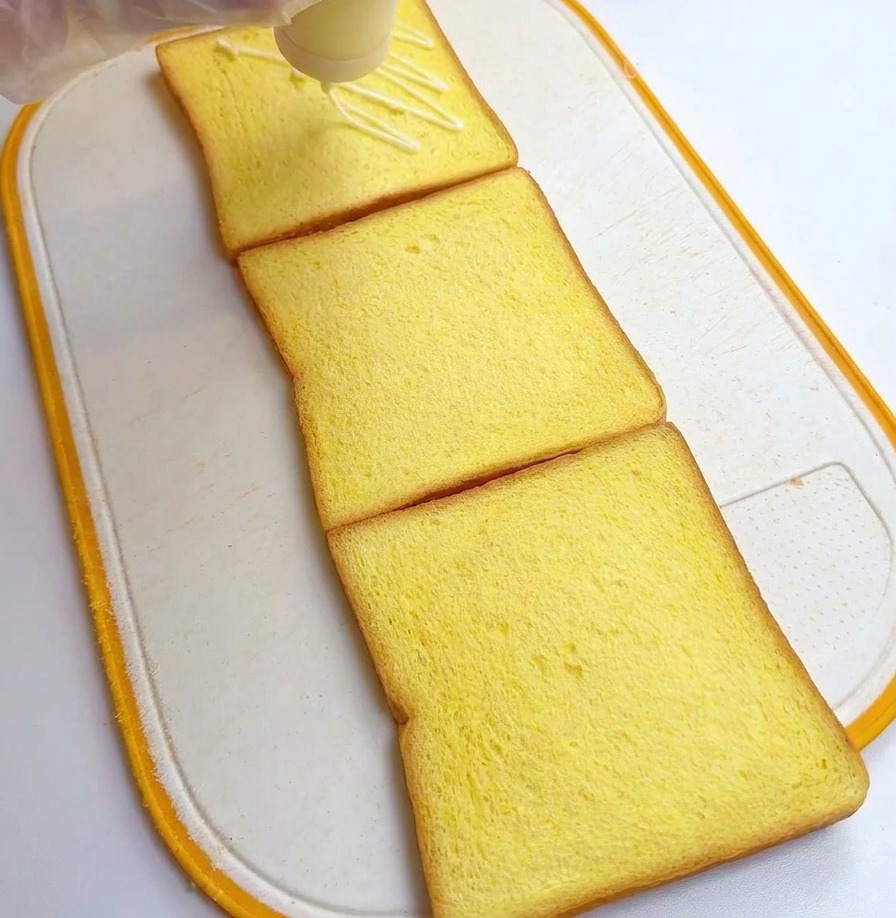 Une personne avec des gants versant de la sauce blanche en zigzag sur trois tranches de pain de mie.