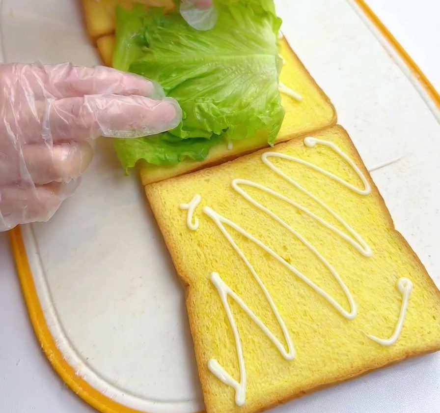 Des mains posant une feuille de laitue verte sur une tranche de pain garnie de sauce.