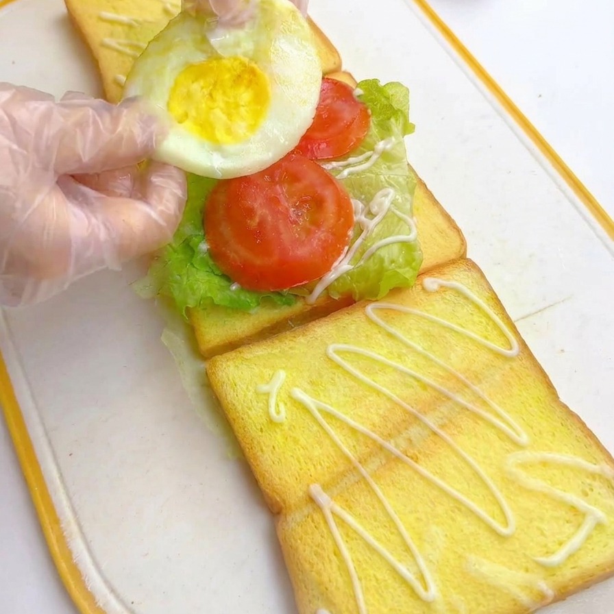 Mains gantées posant un œuf au plat sur les tomates dans le sandwich.