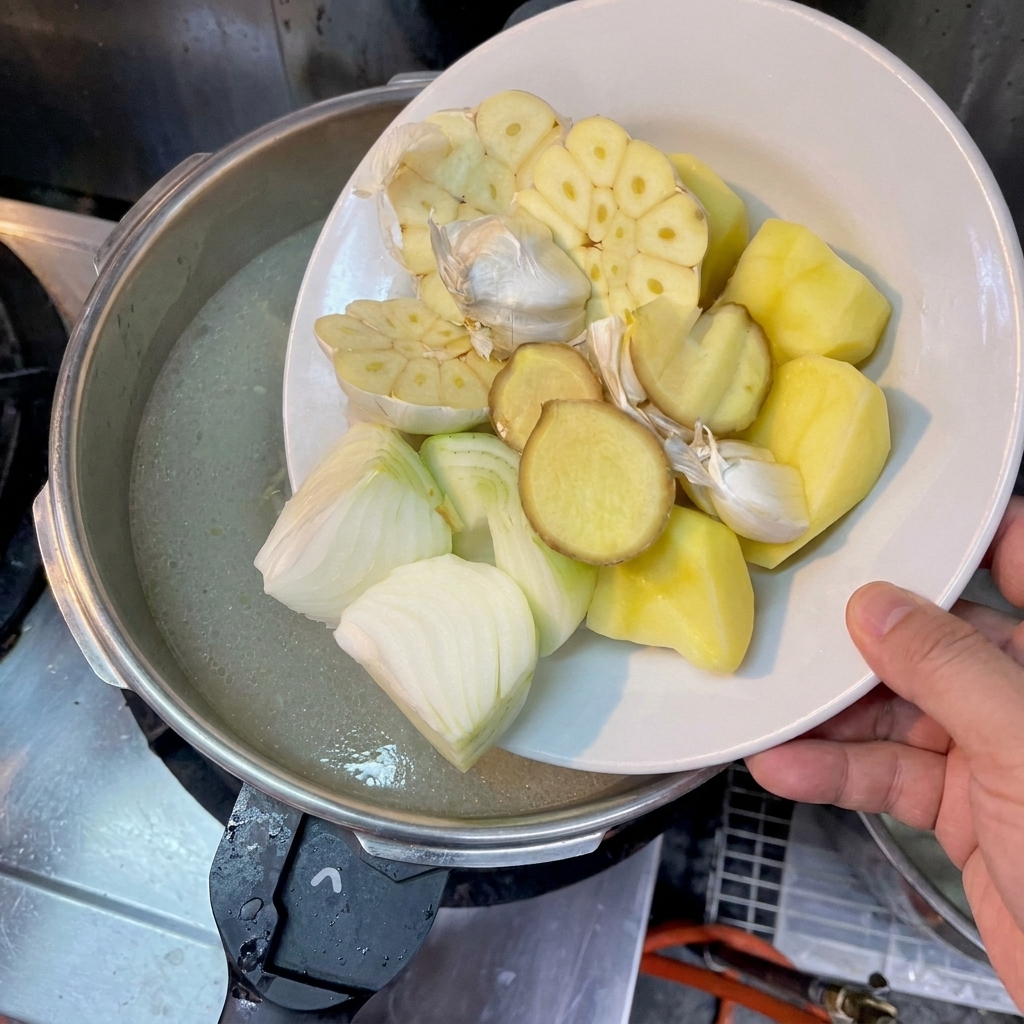 Une main glissant des morceaux de pomme de terre, doignon, de gingembre et dail dans le bouillon.
