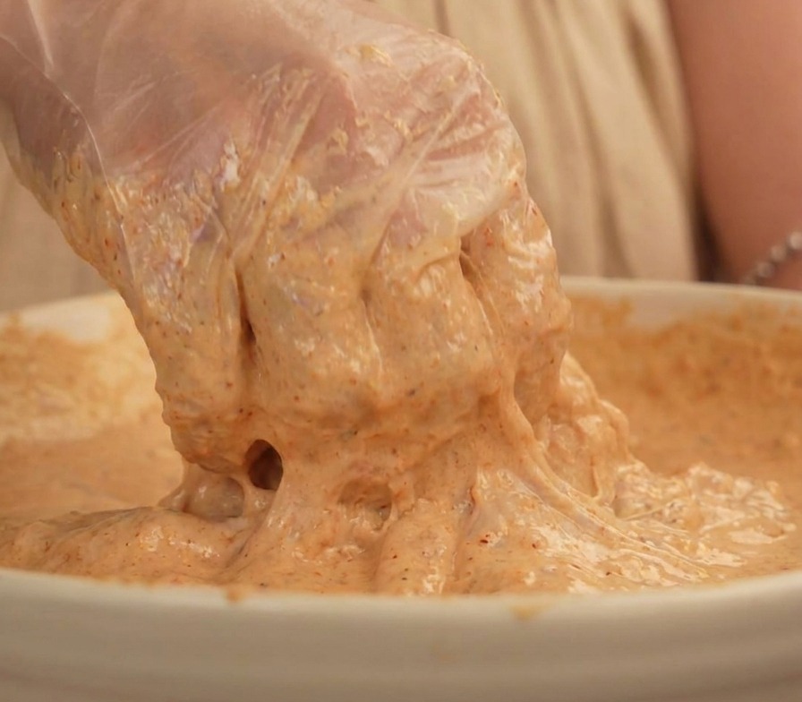 Une main avec un gant plastique soulevant un morceau de poulet bien enrobé de pâte beige épaisse.