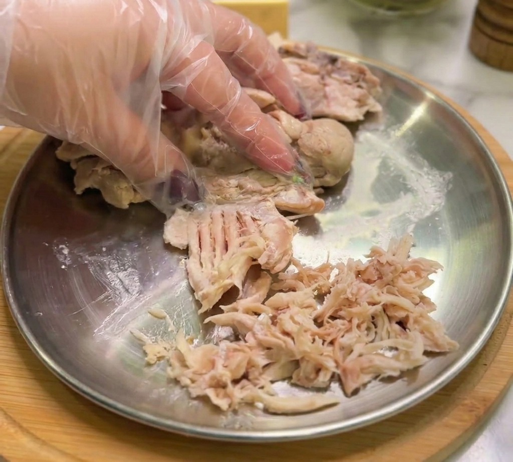 Une main gantée séparant le poulet cuit en fines lanières sur une assiette en métal.