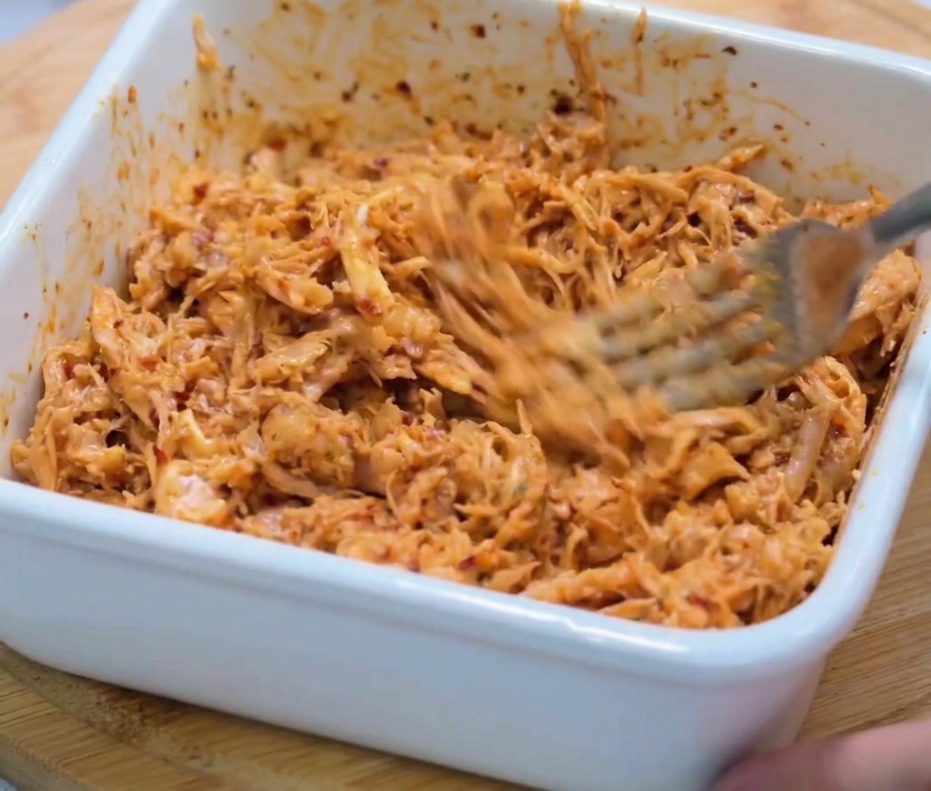 Gros plan du poulet effiloché mélangé à une sauce rouge épaisse avec une fourchette dans un bol blanc.