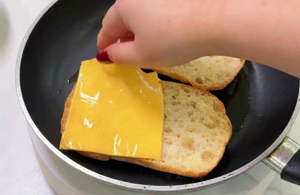 Une main déposant une tranche de fromage fondu sur le pain toasté dans la poêle.