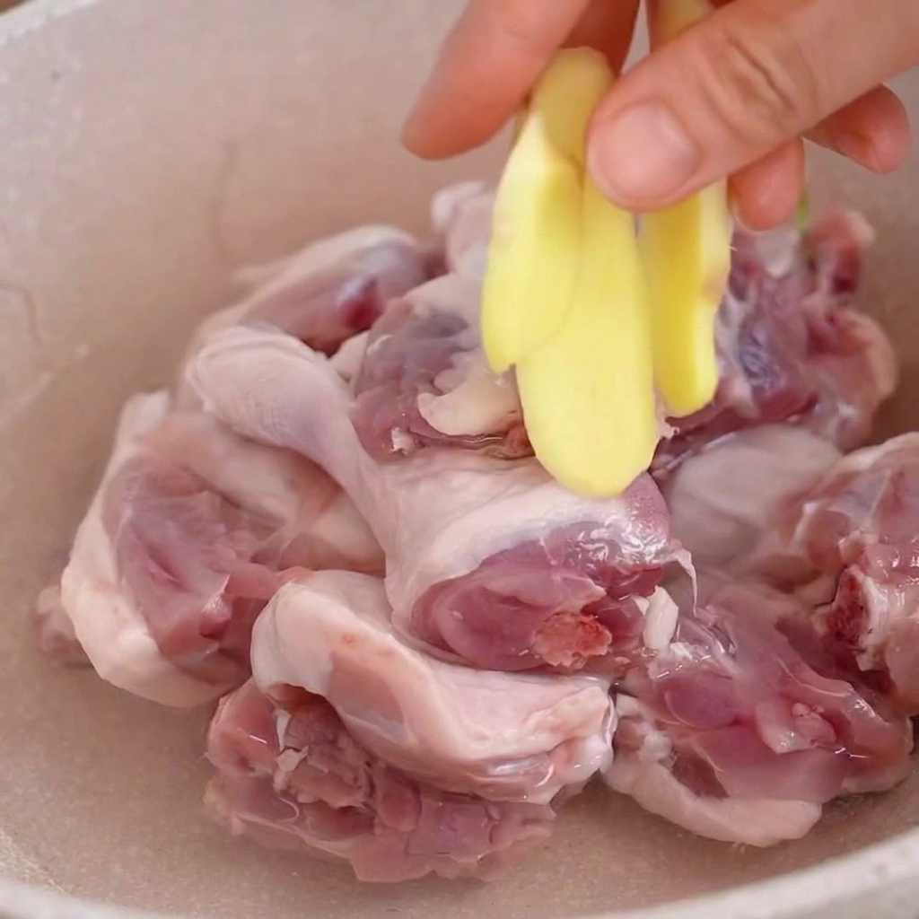 Une main ajoutant des tranches de gingembre frais dans une casserole avec des morceaux de canard crus immergés dans leau.