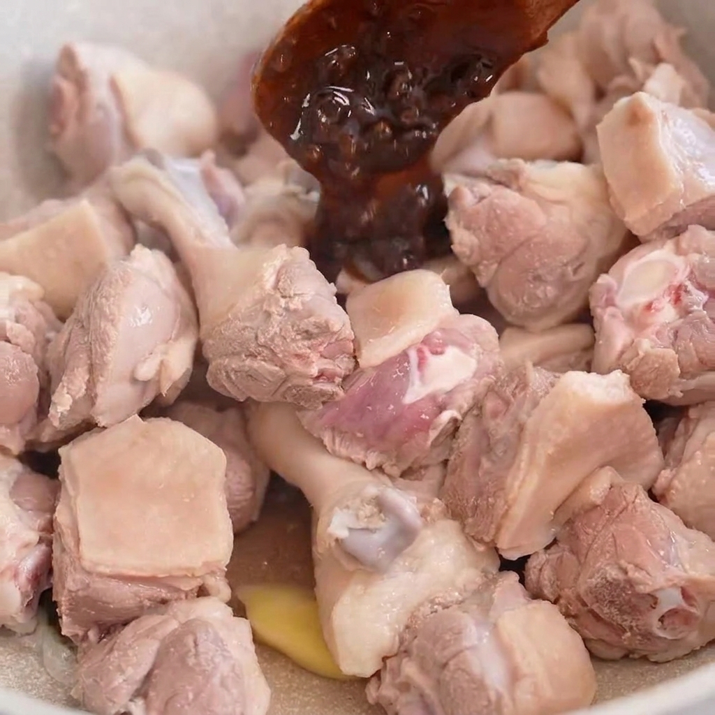 Une cuillère en bois déposant de la pâte de soja foncée sur des morceaux de canard dans une poêle claire.