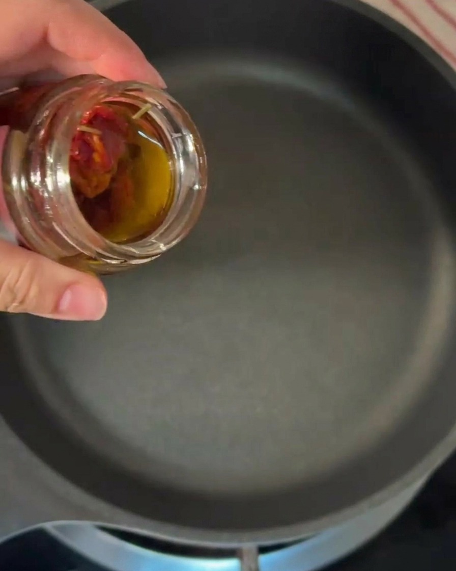 Une main versant de lhuile dolive teintée dorange depuis un bocal en verre dans une poêle noire chaude.