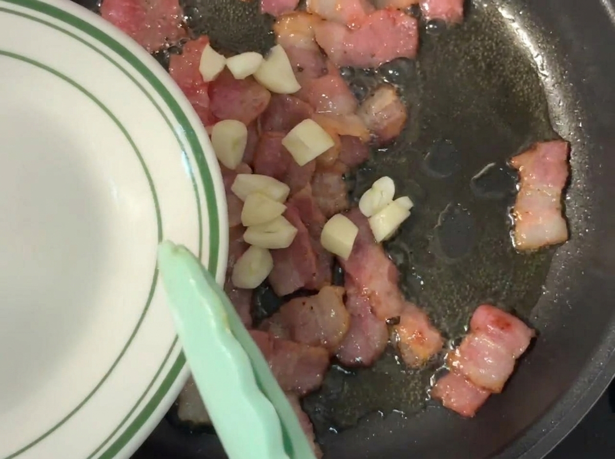 Ail haché ajouté dans une poêle contenant du bacon grésillant et partiellement doré.