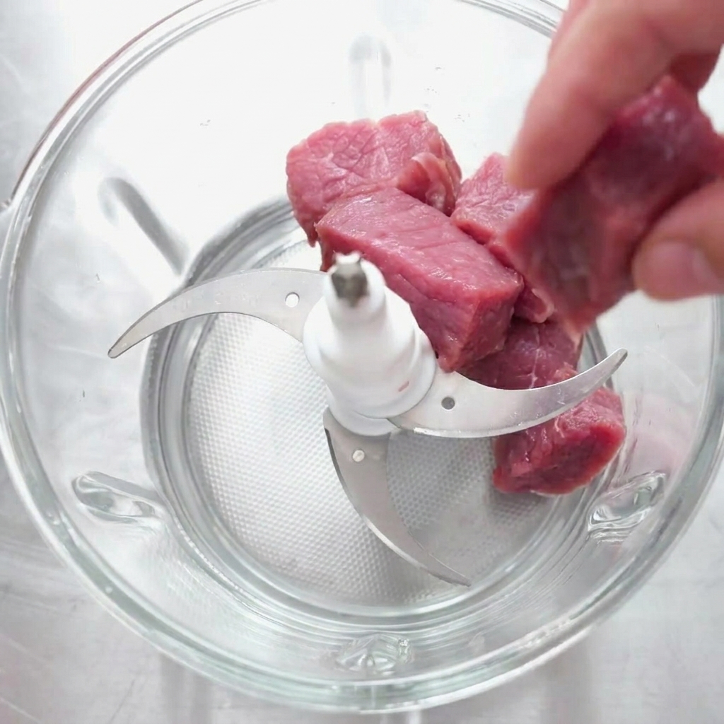 Des morceaux de bœuf cru placés dans le bol en verre dun hachoir électrique équipé de plusieurs lames.