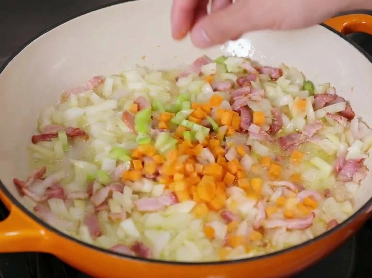 Des carottes en dés ajoutées dans une poêle en fonte contenant des oignons, du céleri et des lardons légèrement rissolés.
