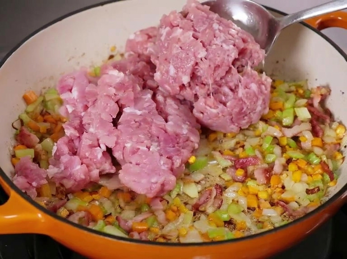 Une grande cuillerée de viande hachée crue ajoutée à une poêle frémissante de légumes en dés et de lardons.