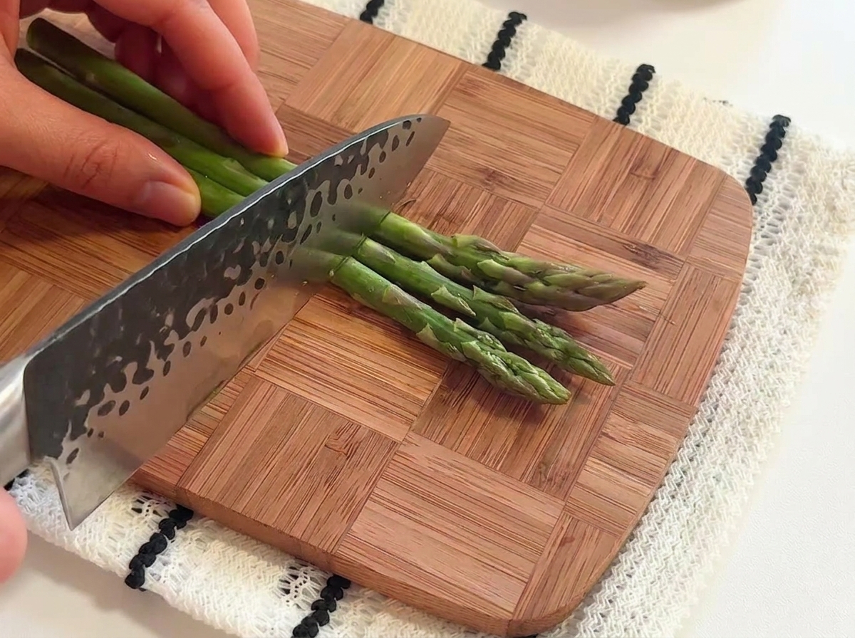 Mains utilisant un couteau de chef pour couper des tiges fraîches dasperges vertes en petits segments sur une planche à découper en bois.