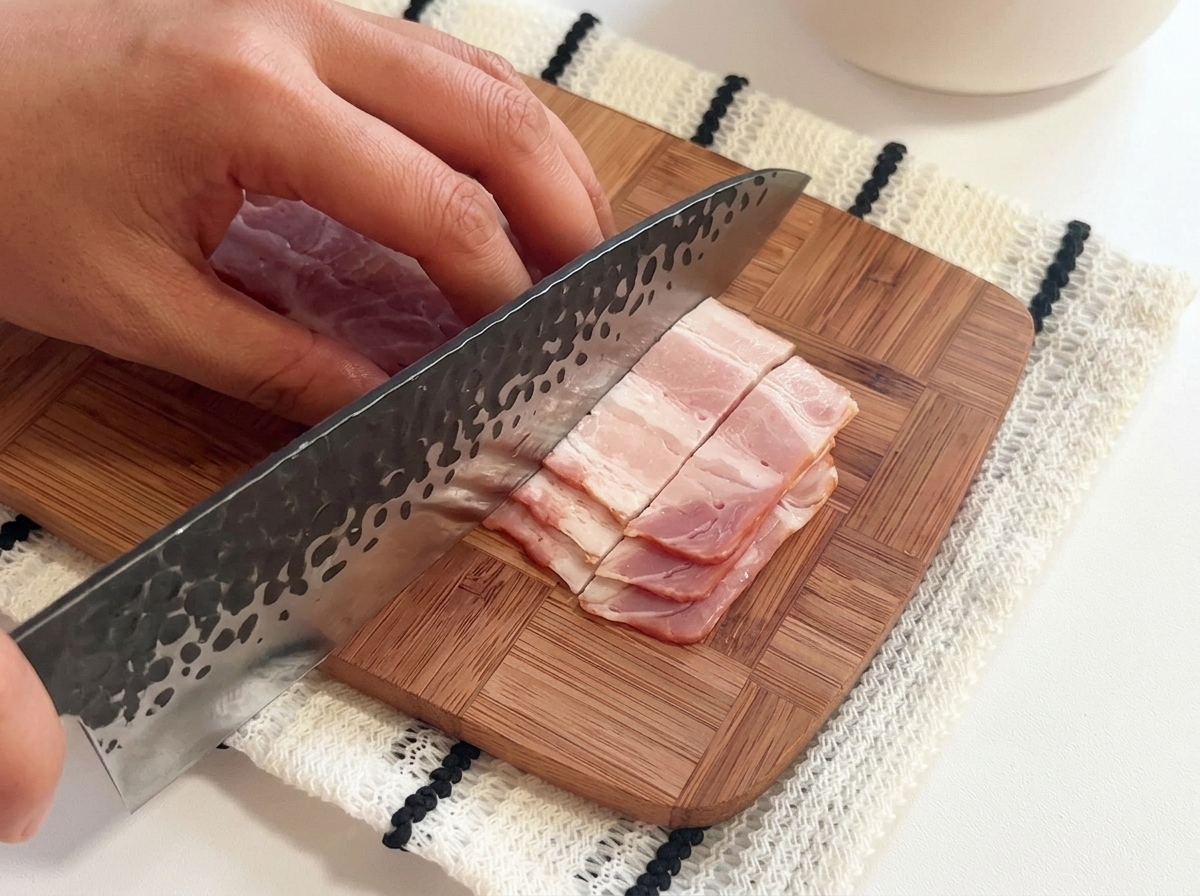Lanières de bacon cru coupées en petits morceaux sur une planche à découper en bois avec un grand couteau.