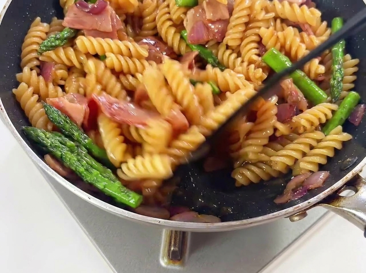 Pâtes, asperges vertes et morceaux de bacon mélangés dans une poêle chaude pour bien les enrober de sauce.