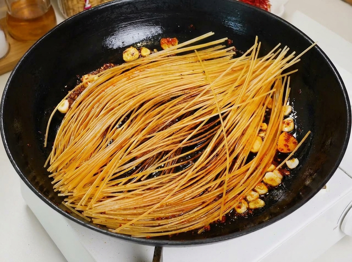 Une louche versant un bouillon de tomate rouge sur des spaghetti grillés dans une poêle noire.