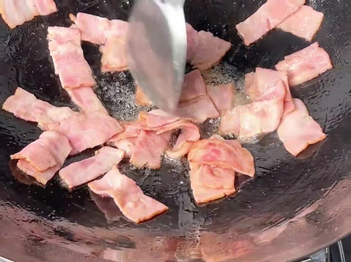 Morceaux de bacon rectangulaires sautés dans un wok en fer avec une spatule en métal.