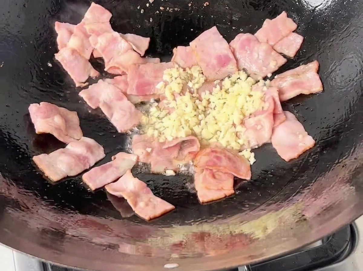 Ail blanc émincé ajouté dans le wok avec le bacon grésillant.