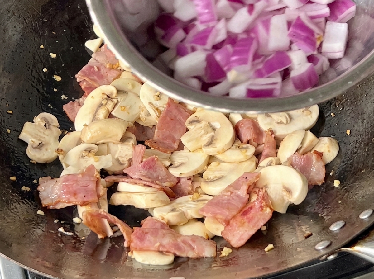 Wok contenant des champignons, du bacon et des oignons rouges émincés en cours de cuisson.