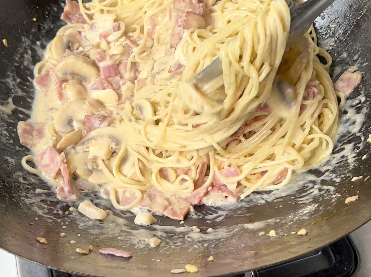 Gros plan de spaghetti mélangés dans la sauce crémeuse à laide de pinces en métal.