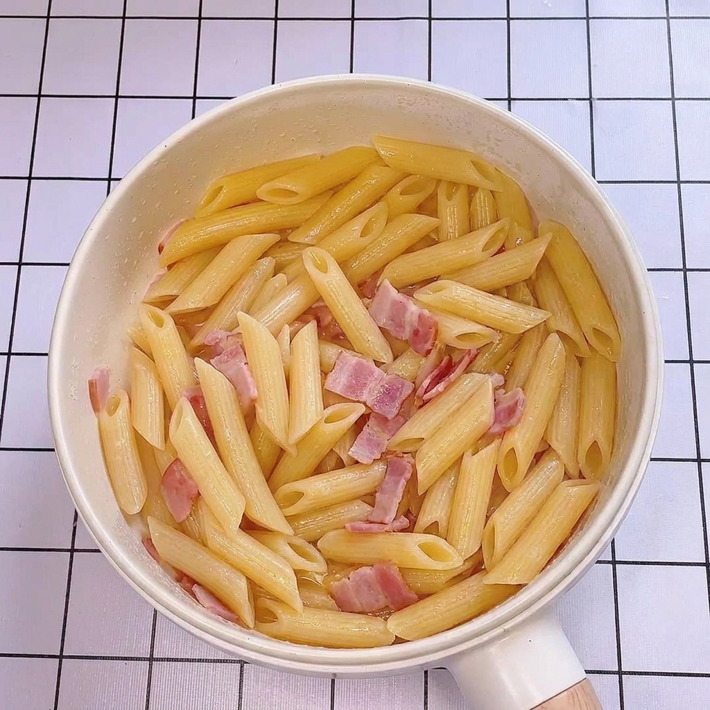 Leau de cuisson des pâtes versée dans la poêle avec les pennes et les lardons, créant un aspect brillant.