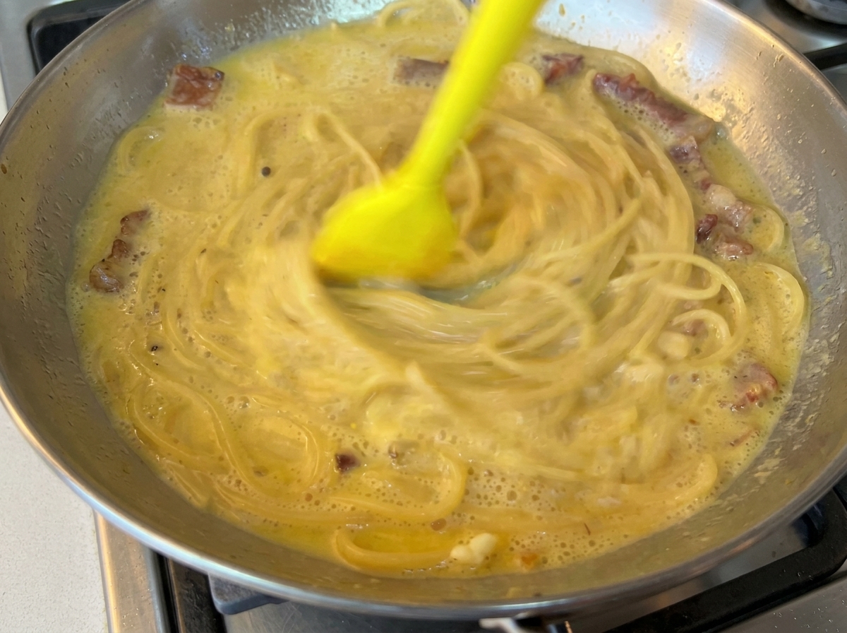 Une spatule en silicone jaune mélangeant rapidement des spaghettis avec une sauce crémeuse aux œufs dans une poêle en inox.