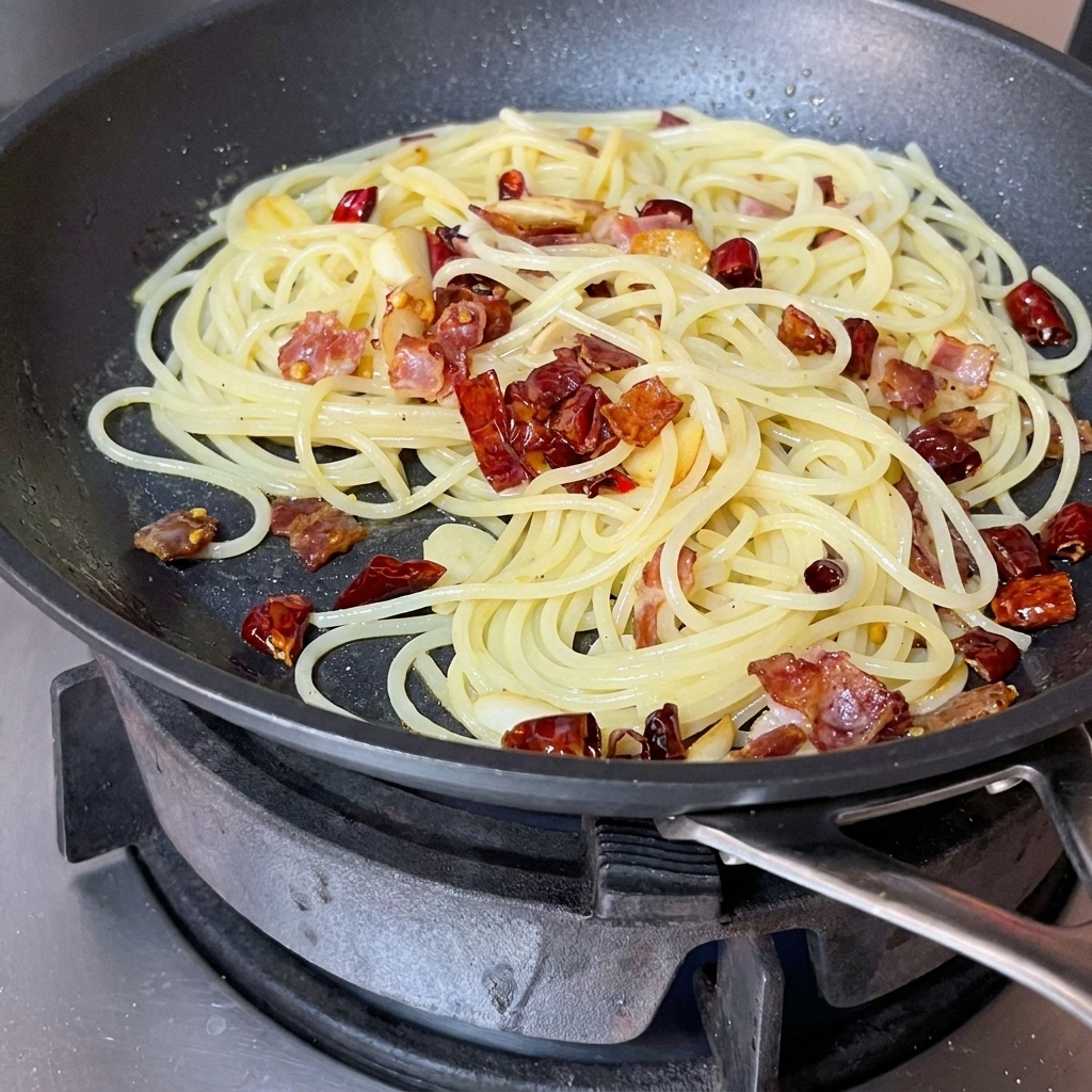 Spaghettis, bacon et aromates mélangés et remués ensemble dans une poêle avec une spatule rouge.