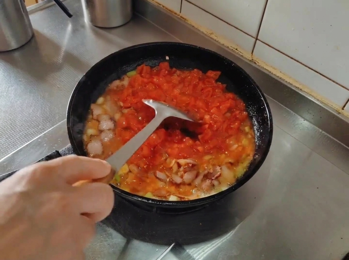 Une spatule en métal écrasant les tomates concassées dans une poêle avec le lard et les aromates.