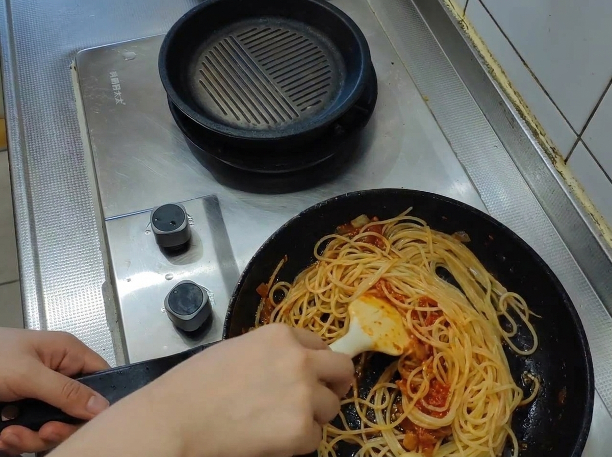 Une louche versant de leau de cuisson bouillante et trouble dans une poêle remplie de spaghetti et de sauce tomate.