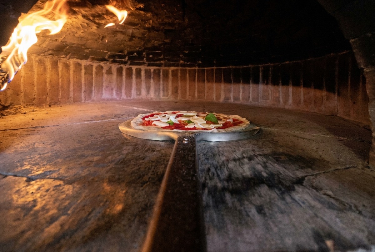 Une pelle à pizza métallique transférant une pizza crue dans un four à bois chaud.