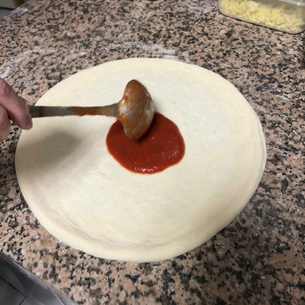 Étaler la sauce tomate rouge sur la pâte à pizza avec des mouvements circulaires à laide dune louche en métal.