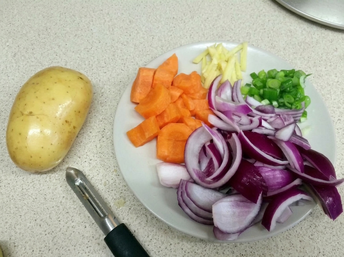 Une assiette blanche contenant des carottes coupées, des oignons rouges, des lamelles de gingembre et des échalotes, à côté dune pomme de terre crue et dun éplucheur.