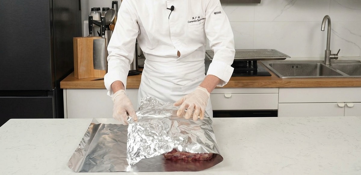 Un chef pliant une grande feuille de papier aluminium argenté sur les côtes de bœuf assaisonnées.