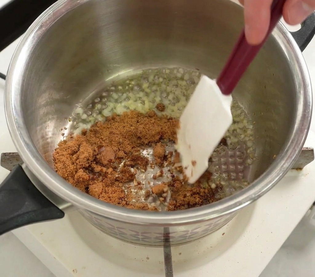 Sucre brun mélangé dans une casserole avec des oignons et de lail à laide dune spatule blanche.