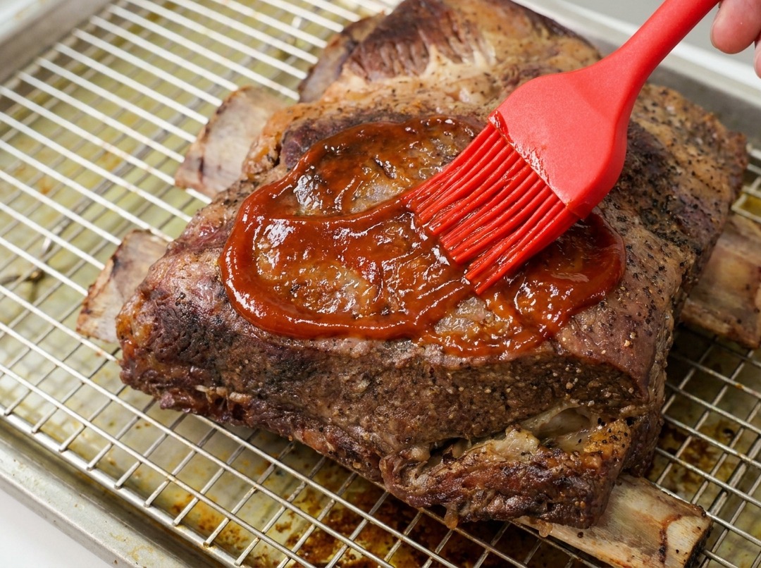 Une main utilisant un pinceau en silicone rouge pour étaler une sauce BBQ épaisse et brillante sur les côtes de bœuf.