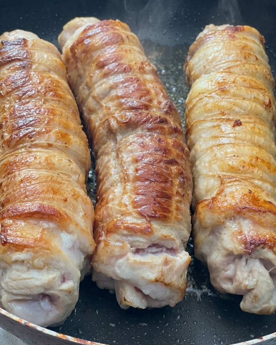 Trois rouleaux de poitrine de porc liés, saisis dans une poêle chaude, montrant une croûte dorée caramélisée.