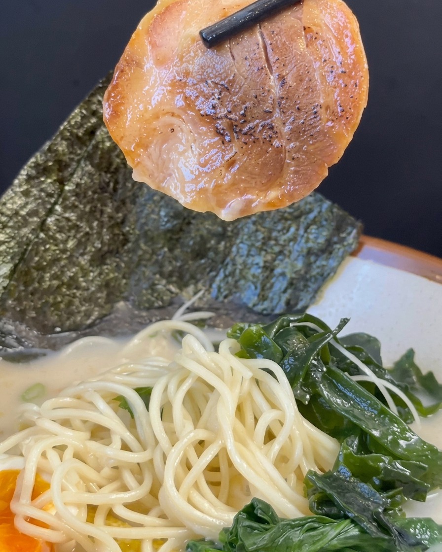 Baguettes noires soulevant une tranche de porc char siu caramélisée au-dessus dun bol de ramen assemblé.