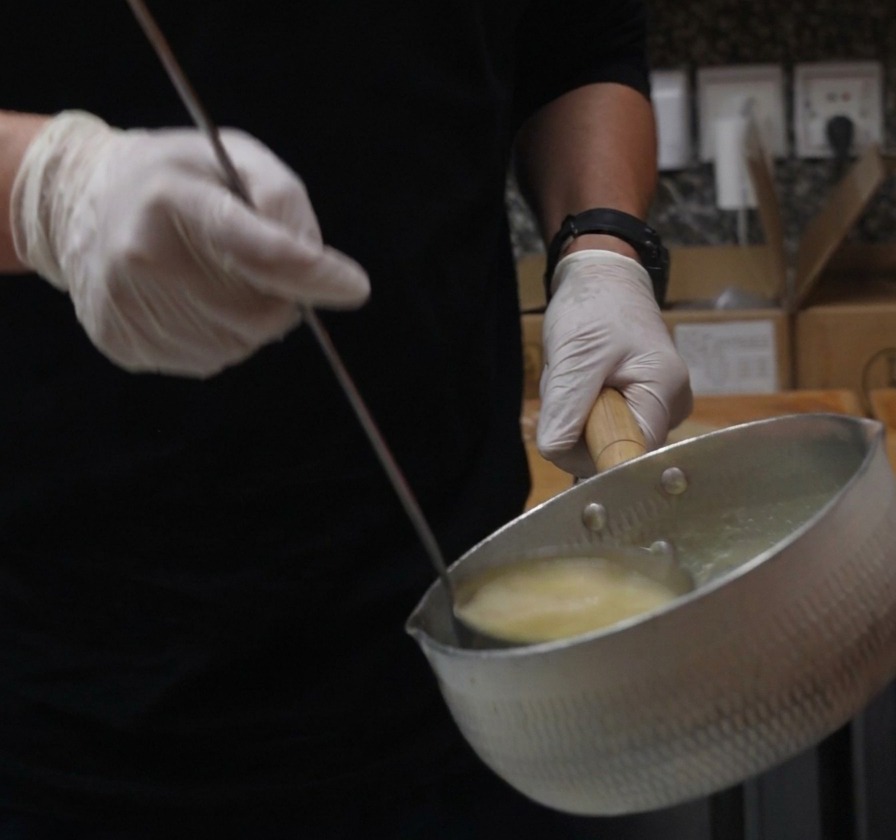 Des mains gantées tenant une casserole en métal et une louche, versant un bouillon paitan de poulet jaune et crémeux.