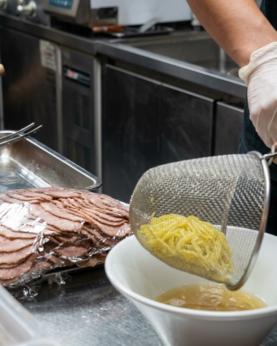 Une passoire métallique déposant des nouilles ramen jaunes fraîchement cuites dans un bol blanc rempli de bouillon crémeux.