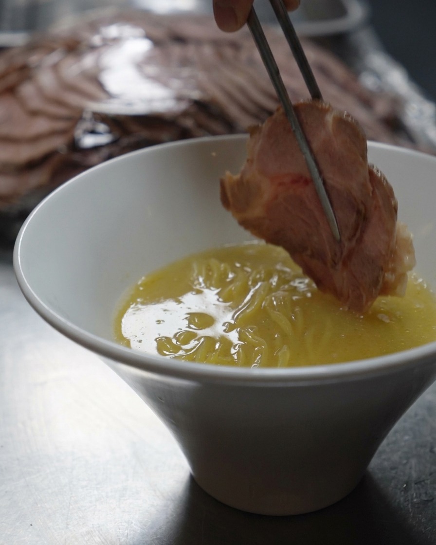 Des baguettes en métal déposant une épaisse tranche de porc saisi dans un bol de nouilles ramen et de bouillon crémeux.