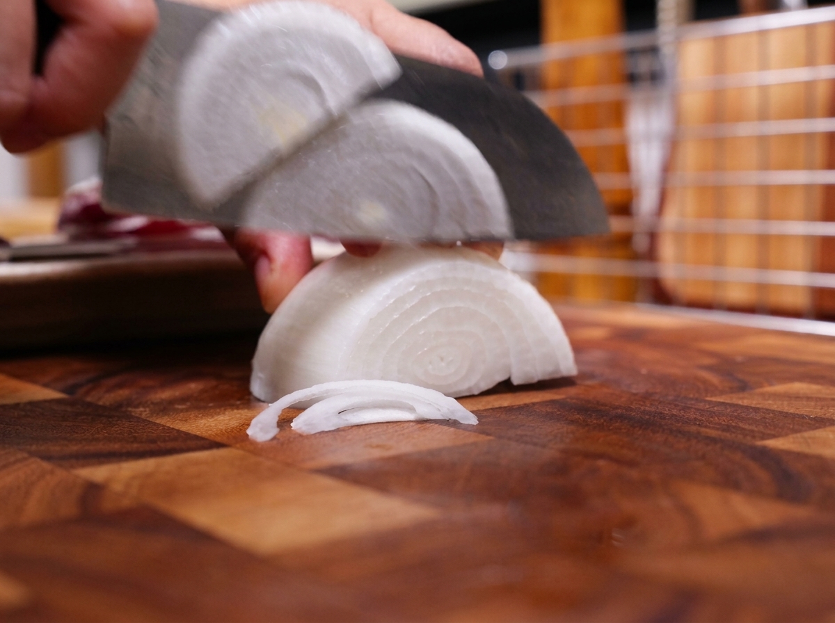 Un couteau de chef coupant la moitié dun oignon blanc en fines lamelles sur une planche à découper en bois.