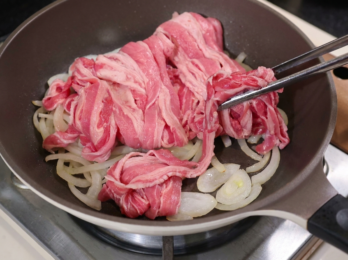 Des pinces en métal déposant de fines tranches de bœuf cru sur les oignons cuits dans une poêle chaude.