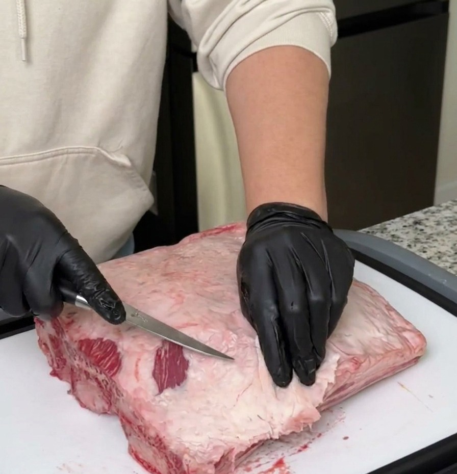 Une personne avec des gants noirs retire lexcès de gras et la membrane argentée dun grand plat de côtes de bœuf.
