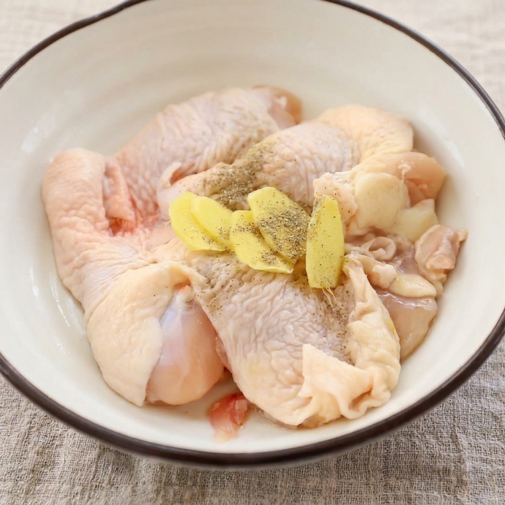 Hauts de cuisse de poulet crus dans un bol, garnis de tranches de gingembre frais et de poivre noir moulu.