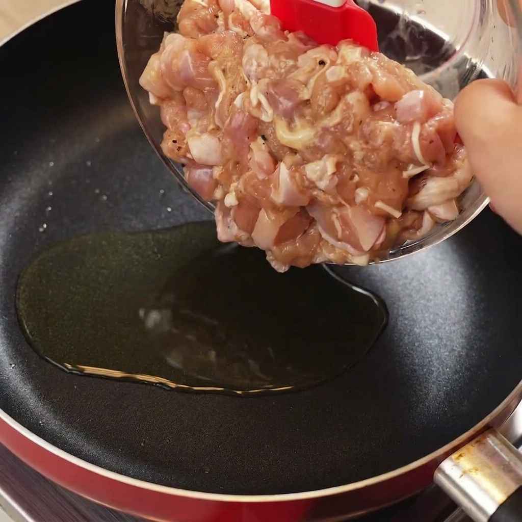 Utilisation dune spatule rouge pour transférer des morceaux de poulet cru mariné dun bol en verre vers une poêle chaude huilée.
