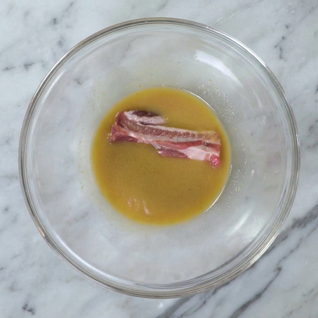 Un travers de porc cru immergé dans la marinade miso lisse à lintérieur dun grand bol en verre.